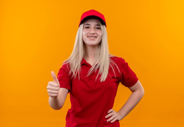 赤いTシャツと歯列矯正器のキャップを身に着けている笑顔の配信の若い女の子は、孤立したオレンジ色の背景に彼女の親指を腰に手を置きます