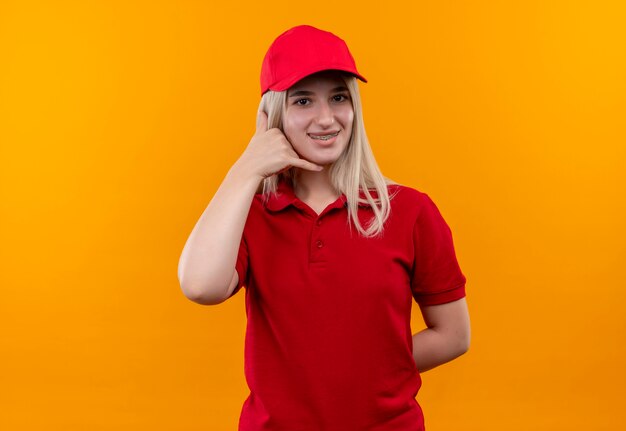 격리 된 오렌지 배경에 전화 제스처를 보여주는 치과 중괄호에 빨간 t- 셔츠와 모자를 입고 배달 어린 소녀 미소