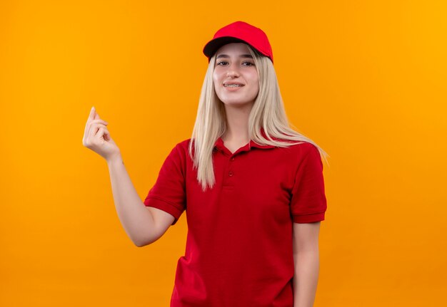 Smiling delivery young girl wearing red t-shirt and cap in dental brace showing tips gesture on isolated orange background