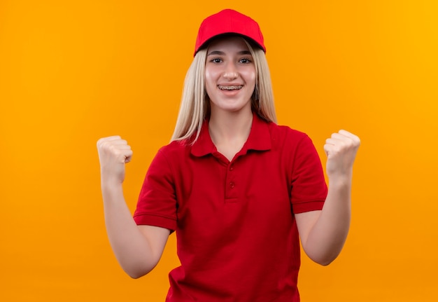孤立したオレンジ色の背景にイエスのジェスチャーを示す歯科ブレースで赤いTシャツとキャップを身に着けている笑顔の配達の若い女の子