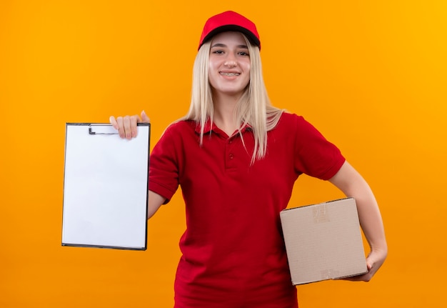 Smiling delivery young girl wearing red t-shirt and cap holding box and clipboard on isolated orange background