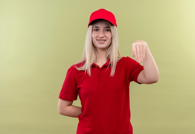 Улыбающаяся девушка из службы доставки в красной футболке в кепке показывает жест на изолированном зеленом фоне