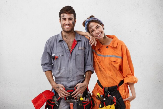 Smiling dirty woman leans on shoulder of man mechanic, helps him to repair car on work station