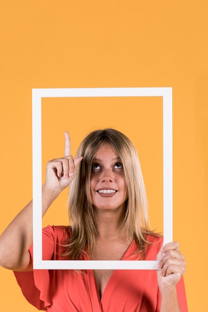 Smiling disable woman holding frame cutout with pointing upward on plain surface