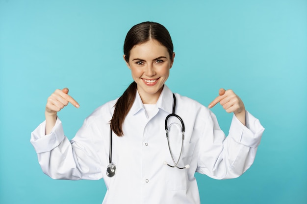 Smiling doctor medical worker pointing fingers at logo clinic banner showing advertisement wearing w...