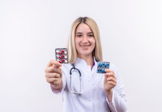 Бесплатное фото Улыбаясь доктор молодая блондинка носить стетоскоп и медицинский халат в стоматологической скобе, держа таблетки обеими руками на изолированном белом фоне
