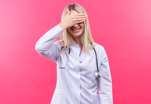 Улыбающаяся молодая блондинка со стетоскопом в медицинском халате и стоматологической скобе закрыла глаза на изолированном розовом фоне