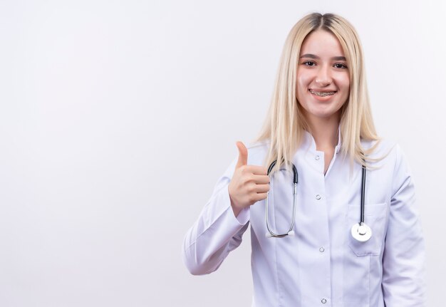 Smiling doctor young blonde girl wearing stethoscope and medical gown in dental brace her thumb up on isolated white background