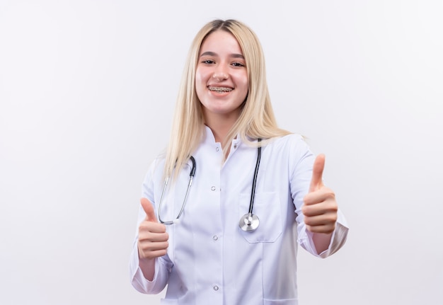 Улыбаясь доктор молодая блондинка носить стетоскоп и медицинский халат в стоматологической скобке ее пальцы на изолированном белом фоне