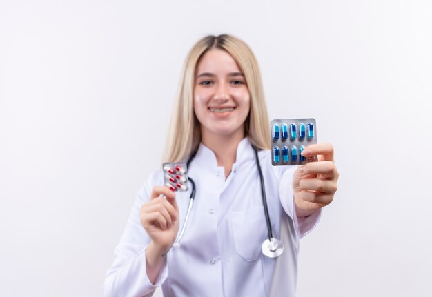 Smiling doctor young blonde girl wearing stethoscope and medical gown in dental brace holding pills with both hands on isolated white background