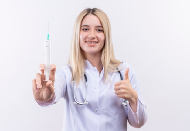 Улыбаясь доктор молодая блондинка со стетоскопом и медицинским халатом в стоматологической скобе, держа шприц вверх большим пальцем на изолированном белом фоне