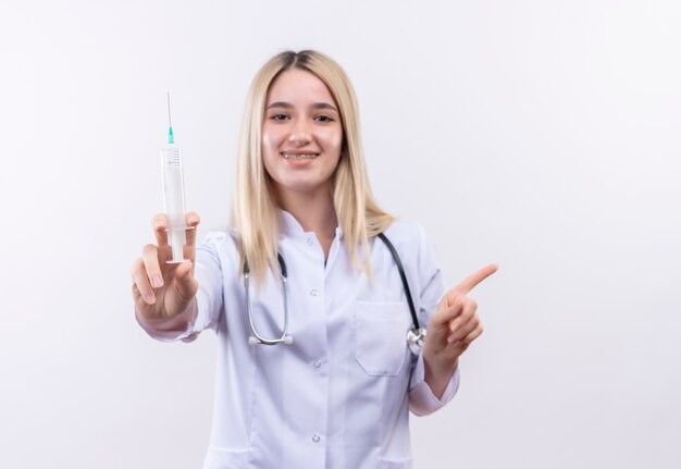 Smiling doctor young blonde girl wearing stethoscope and medical gown in dental brace holding syringe points to side on isolated white background