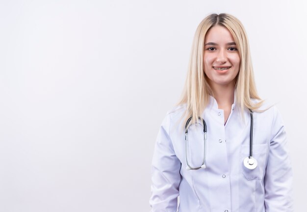 Smiling doctor young blonde girl wearing stethoscope and medical gown in dental brace on isolated white background