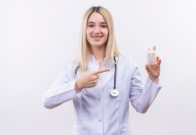 Smiling doctor young blonde girl wearing stethoscope and medical gown in dental brace points to empty can holding on her hand on isolated white background