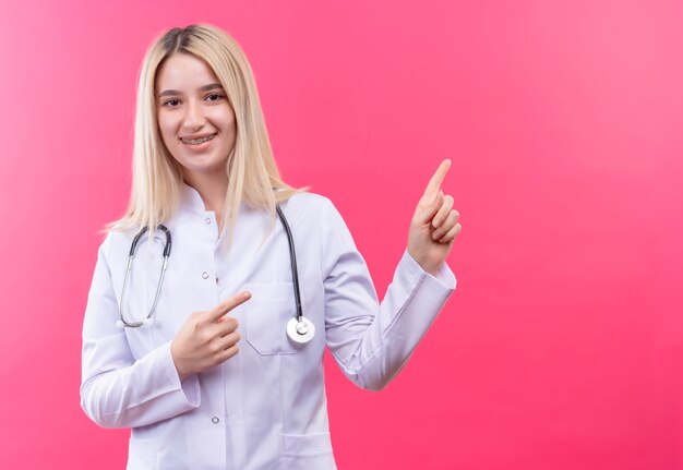医療用ガウンと歯列矯正器で聴診器を身に着けている笑顔の医者の若いブロンドの女の子は、孤立したピンクの背景に両手で横を指します