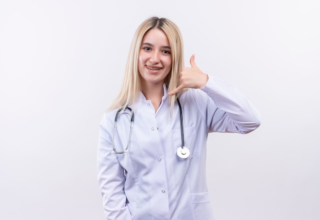 Smiling doctor young blonde girl wearing stethoscope and medical gown in dental brace showing call gesture on isolated white background