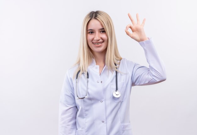Smiling doctor young blonde girl wearing stethoscope and medical gown in dental brace showing okay gesture on isolated white background
