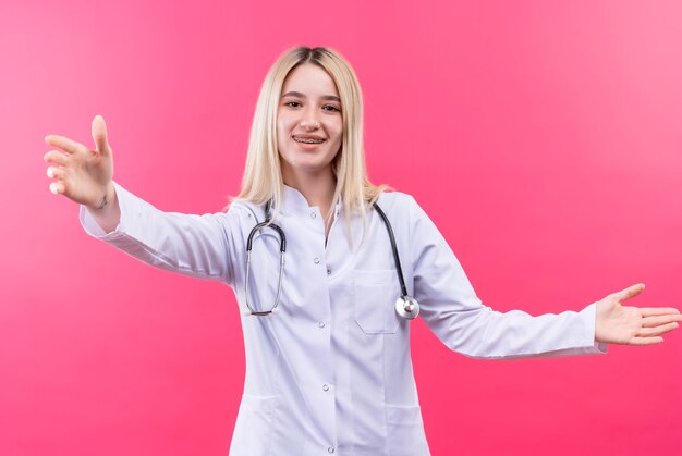 Smiling doctor young blonde girl wearing stethoscope in medical gown and dental brace showing size on isolated pink background