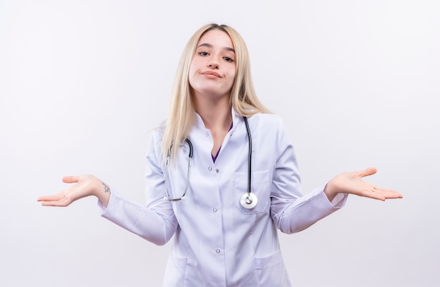 청진 기 및 격리 된 흰색 배경에 어떤 제스처를 보여주는 의료 가운을 입고 웃는 의사 젊은 금발 소녀