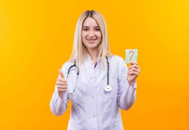 Улыбающаяся молодая девушка-врач со стетоскопом в медицинском халате и стоматологической скобе держит бумажный вопросительный знак большим пальцем на изолированном желтом фоне