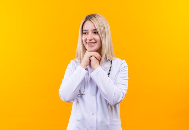 Улыбающаяся молодая девушка-врач со стетоскопом в медицинском халате и стоматологической скобе положила обе руки под подбородок на изолированном желтом фоне
