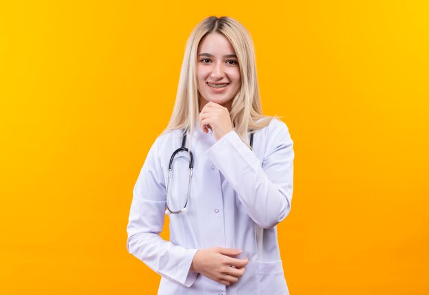 Улыбающаяся молодая девушка-врач со стетоскопом в медицинском халате и стоматологической скобе положила руку на подбородок на изолированном желтом фоне