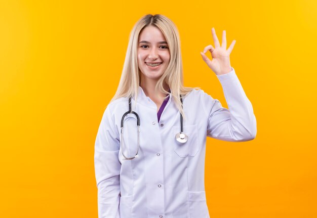 Улыбающаяся молодая девушка-врач со стетоскопом в медицинском халате и стоматологической скобе показывает нормальный жест на изолированном желтом фоне