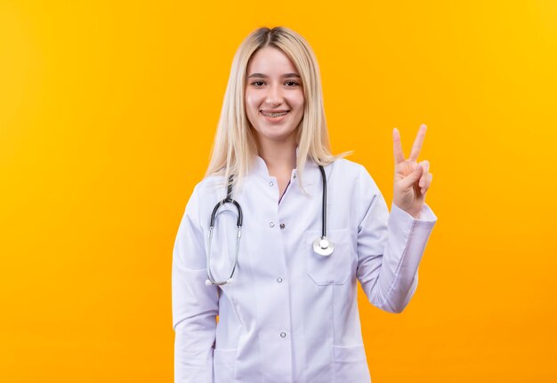 Smiling doctor young girl wearing stethoscope in medical gown and dental brace showing peace gesture on isolated yellow background