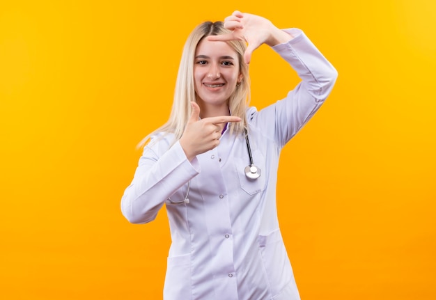 Улыбающаяся молодая девушка-врач со стетоскопом в медицинском халате и стоматологической скобе, показывающая жест фото на изолированном желтом фоне