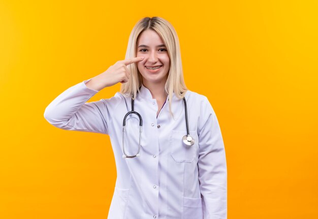 Smiling doctor young girl wearing stethoscope in medical gown and dental braces points to eye on isolated yellow background