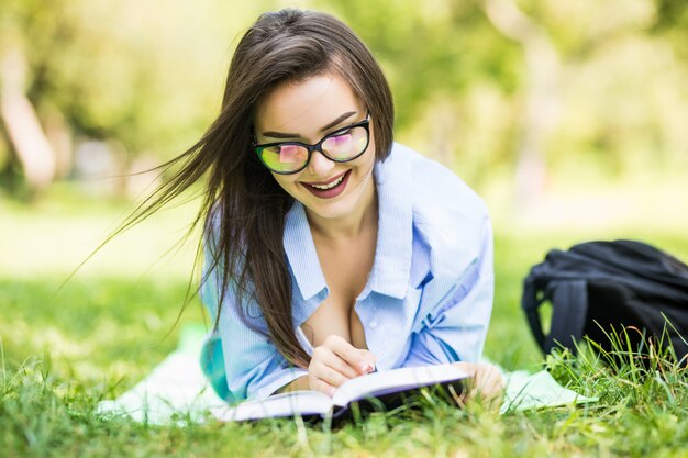 Smiling dreamy teen girl lying on grass in park with notebook