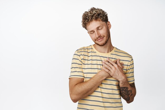 Smiling dreamy young man close his eyes holds hands on chest and daydreaming imaging something lovely and heartwarming cherish memories white background