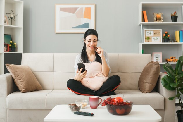 Smiling eat biscuits young girl with pillow sitting on sofa behind coffee table holding and looking at phone in living room