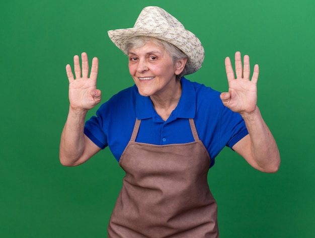 Smiling elderly female gardener wearing gardening hat gesturing four with fingers isolated on green wall with copy space