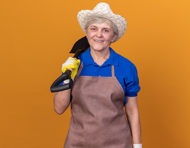 Sorridente giardiniere donna anziana che indossa cappello e guanti da giardinaggio che tengono la vanga sulla spalla isolata sulla parete arancione con spazio per le copie