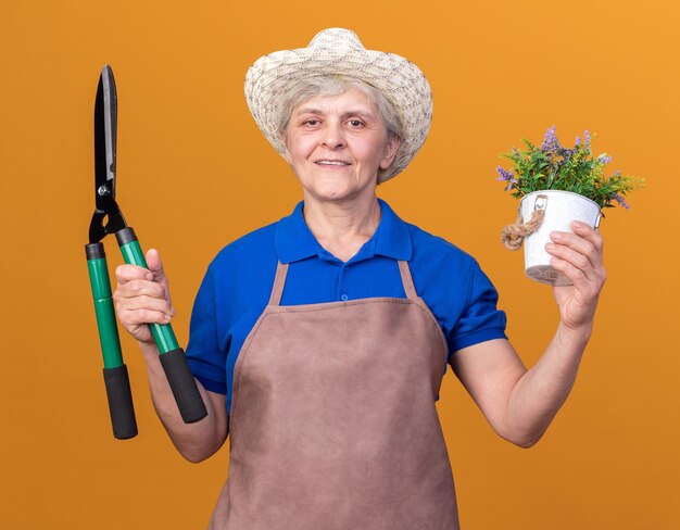 オレンジ色の園芸はさみと植木鉢を保持している園芸帽子を身に着けている年配の女性の庭師の笑顔