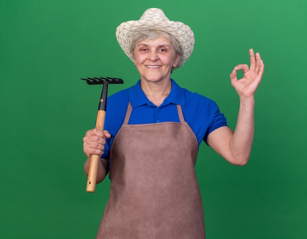Foto gratuita sorridente giardiniere donna anziana che indossa cappello da giardinaggio tenendo rastrello e gesticolando segno ok