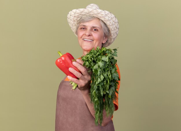 Smiling elderly female gardener wearing gardening hat holding red peppers and bunch of coriander isolated on olive green wall with copy space