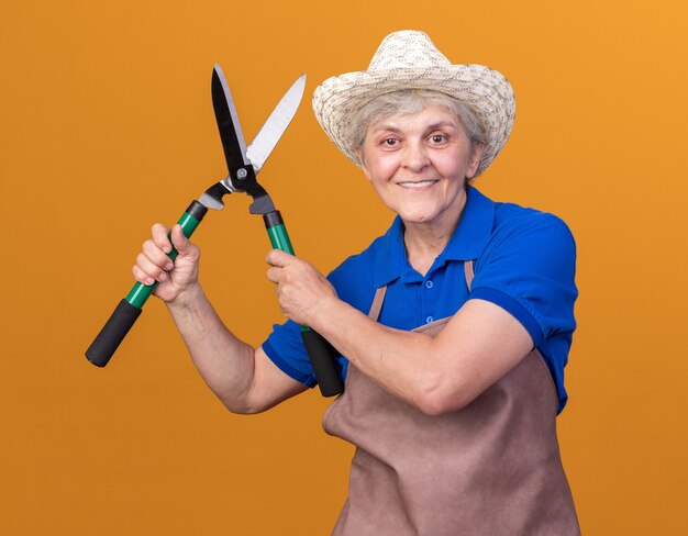 원예 모자를 쓰고 웃는 노인 여성 정원사는 오렌지 원예 가위를 보유하고 있습니다.