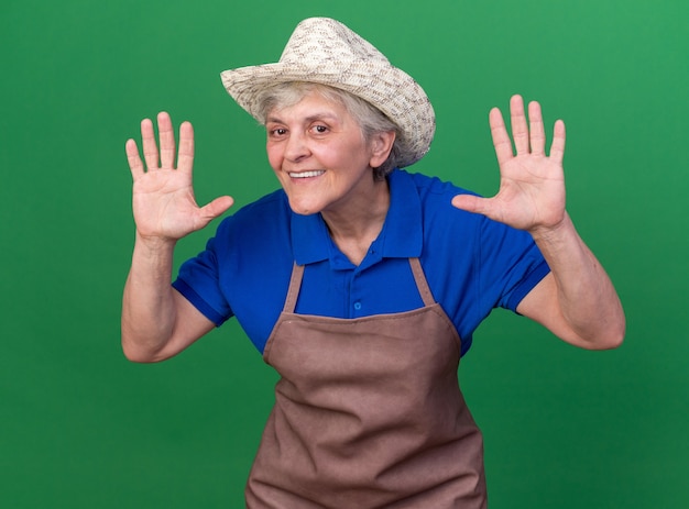 Smiling elderly female gardener wearing gardening hat standing with raised hands isolated on green wall with copy space