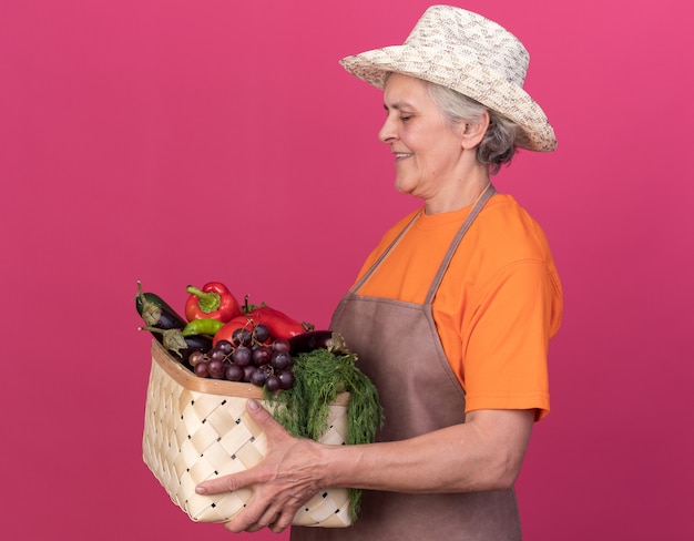 ガーデニング帽子をかぶって笑顔の年配の女性庭師は、野菜のバスケットを持って見て横に立っています