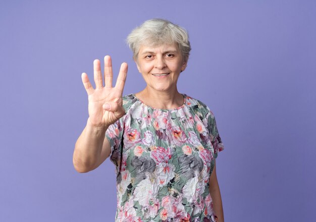 Smiling elderly woman gestures four with hand isolated on purple wall