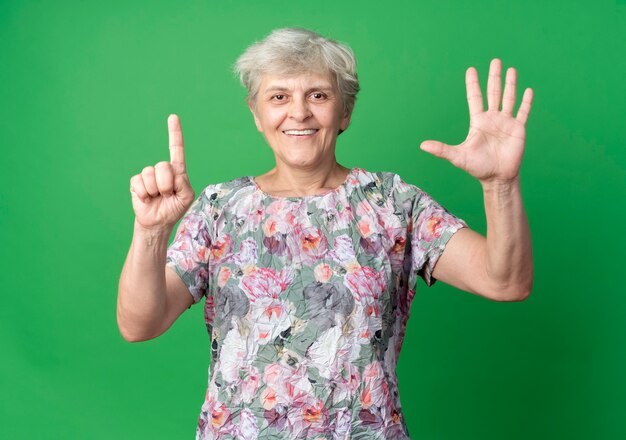 Smiling elderly woman gestures six with hands isolated on green wall