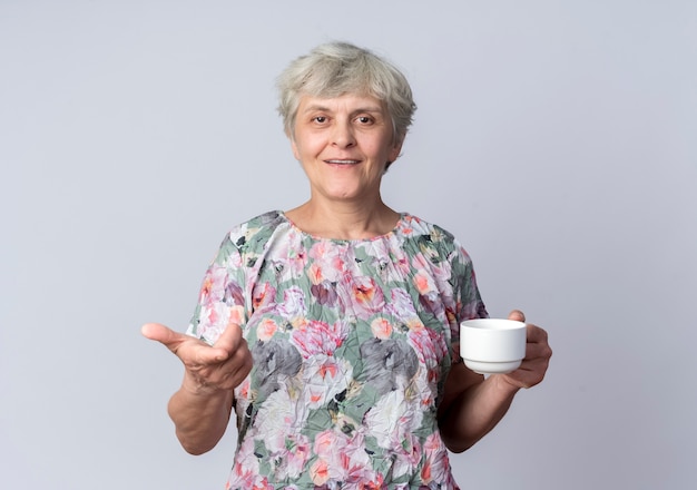La donna anziana sorridente tiene la tazza e punti isolati sulla parete bianca