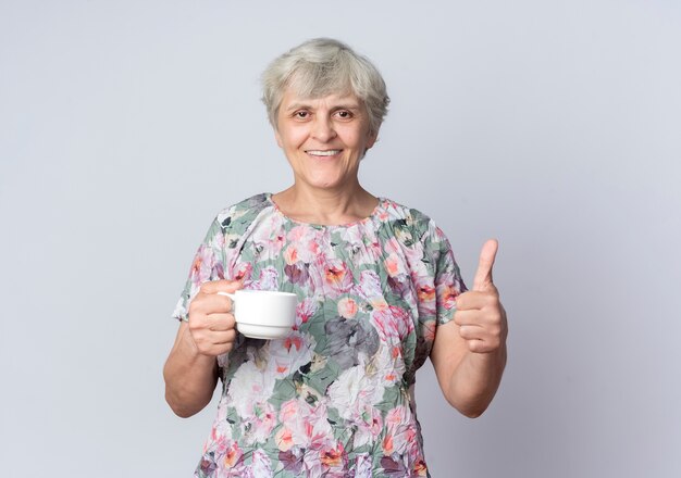 Smiling elderly woman holds cup and thumbs up isolated on white wall