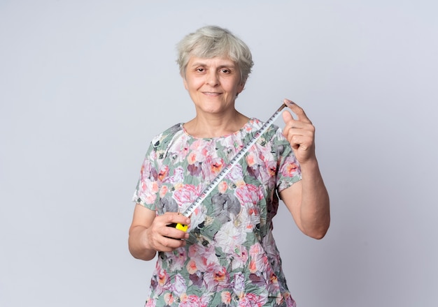 Smiling elderly woman holds tape measure isolated on white wall