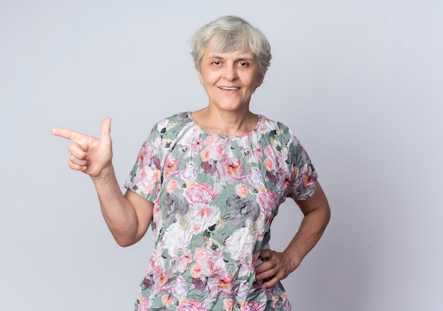 Smiling elderly woman puts hand on waist pointing at side isolated on white wall