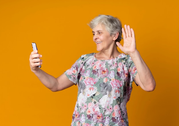 Улыбающаяся пожилая женщина поднимает руку, держа и глядя на телефон, изолированный на оранжевой стене