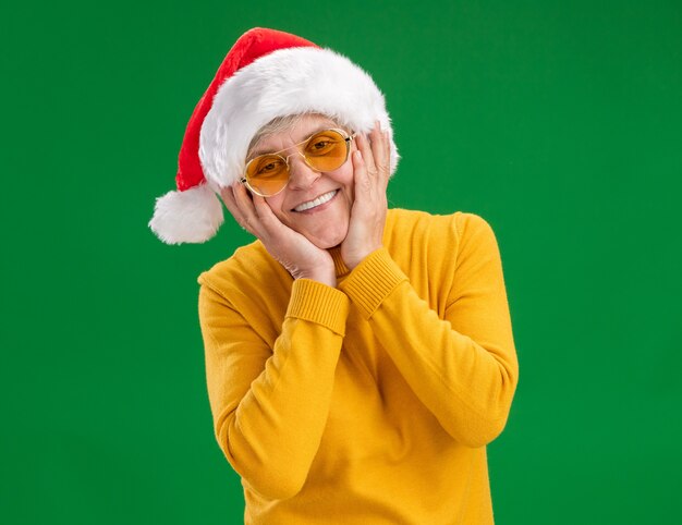 Smiling elderly woman in sun glasses with santa hat puts hands on face isolated on green background with copy space