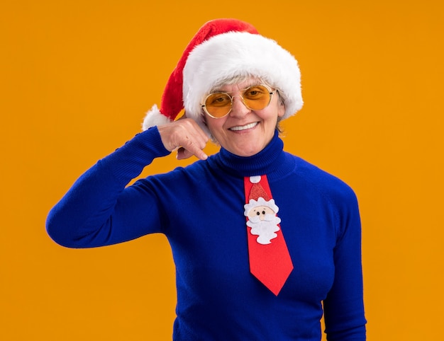 smiling elderly woman in sun glasses with santa hat and santa tie gesturing call me sign isolated on orange background with copy space